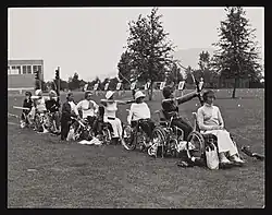 Carte postale en noir et blanc. Une rangée de femmes en jupe pratiquent le tir à l'arc, assises sur leurs fauteuils roulants.