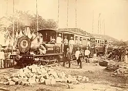 Photo ancienne d'une locomotive à l'arrêt, dans laquelle se trouve un groupe de personnes, dont de nombreux hommes coiffés de casques coloniaux.