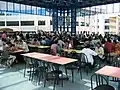 Grande et haute salle vitrée, à l’étage, tables de couleurs différentes selon les rangées et chaises.
