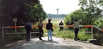 Photographie de la fin d'une route forestière marquée par une barrière horizontale. Quatre personnes dont deux en uniforme sont debout près de la barrière. Un autre homme en uniforme se trouve un peu plus loin dans un champ. À l'arrière-plan, on distingue une clôture métallique et une tour de surveillance avec un sommet octogonal.