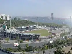 Photographie d'un stade de football, dans son ensemble, avec la route au premier plan et la mer en arrière-plan.