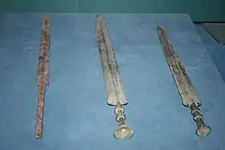 Photographie de deux épées en bronze et d'une épée en fer de tailles similaires. Celle de gauche se distingue des deux autres par la forme de sa lame et de son manche.