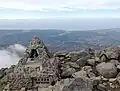 Photo couleur du bord d'un sommet de montagne. Des rochers gris parsèment le sol de terre marron. En bas, à gauche : un monument pyramidal en ciment incrusté de pierre et à l'intérieur évidé. Au deuxième plan : une plaine ; un ciel nuageux, en arrière-plan.