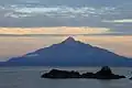 Photo couleur d'un volcan, sur une île, depuis une île voisine, sous un ciel nuageux peu lumineux.