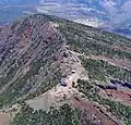 Photo couleur montrant le bord du cratère d'un volcan vu du ciel. Les pentes sont boisées et exposent des bandes de terre à nu par endroits. Trois bâtiments de petite taille sont visibles sur l'arête du cratère.