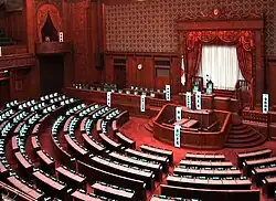 Salle de grande taille à la décoration opulente de bois sculpté. Une longue tribune avec deux rangées de bureaux occupe le mur du fond, les bureaux et fauteuils des conseillers formant un hémicycle} devant elle.