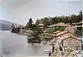 Photo peinte de bâtiments en bois sur la rive (à droite) d'une étendue d'eau, un ciel nuageux en arrière-plan.