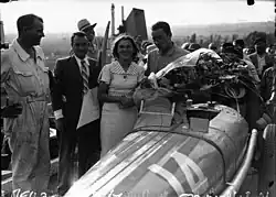 Photo d'un homme en tenue de pilote tenant un bouquet de fleur et félicité par des hommes en costume.