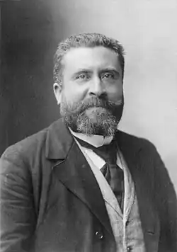 Photo noir et blanc du buste d'un homme barbu portant manteau, veston et cravate.