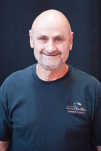 Photographie couleur d’un homme, de tronc, chauve, souriant, avec une barbe de 3&nbsp;jours blanche et un t-shirt bleu marine