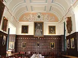Un hall lambrissé de bois, avec un grand portrait d'Elizabeth I au milieu du mur du fond, d'autres portraits sur les murs ; un plafond de plâtre blanc à décor bleu clair
