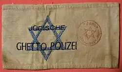 Brassard en tissu beige sur lequel est cousue une étoile de David bleue et est portée la mention « Jüdische Ghetto Polizei » en lettres capitales noires.