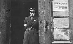 Un policier debout se tient dans l’embrasure d’une porte. À sa droite, une plaque sur le mur extérieur indique (en allemand puis en polonais) : « Service d’ordre du Judenrat de Varsovie, gestion du quatrième arrondissement ».