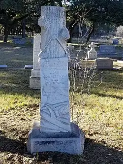 stèle de tombe, sculptée en forme de tronc d'arbre étêté auquel est accroché une bannière portant l'inscription tombale.