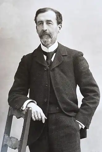 Portrait photographique noir et blanc d’un homme barbu en pied, portant costume.