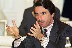Photographie d'un homme brun et moustachu, il est assis et tient un discours.