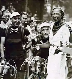 Photographie de presse en noir et blanc montrant l'actrice à droite, se tenant debout à côté des deux coureurs cyclistes avant le départ de la course.