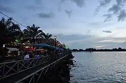 Photographie d'une promenade le long de la mer au crépuscule. De nombreux luminaires sont allumés et plusieurs personnes sont attablées à divers restaurants.