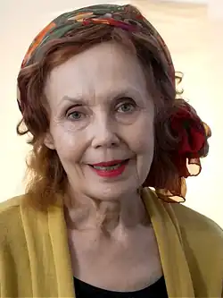 Phtographie en couleur du visage d'une femme âgée portant un foulard rouge sur le crâne et des vêtements jaune et noir