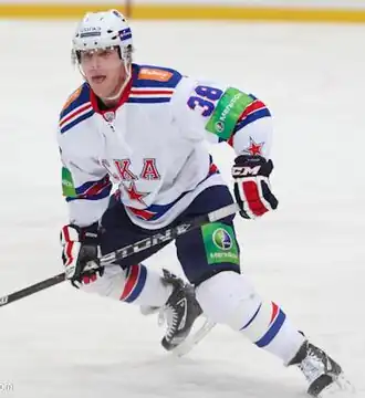 Photographie couleur d’un hockeyeur en pied, les jambes pliées, de trois-quarts face, tirant la langue