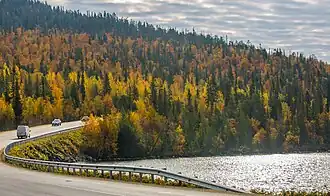 Vue d'une route à gauche longeant la rive d'un lac, avec en second plan une colline recouverte de taïga aux couleurs automnales.