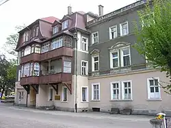 Bâtiment de quatre étages. Les murs du premier sont de couleur beige, et le reste est gris. Le toit est brun. Le bâtiment a deux balcons en bois sur sa face.