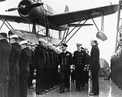 Photographie de plusieurs officiers passant en revue des marins. Un hydravion se trouve à l’arrière-plan.