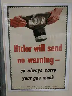 Photo en couleurs d'une affiche publiée par le ministère de la Sécurité civile anglais pendant la Seconde Guerre mondiale. Sur un fond vert foncé, un rectangle de couleur blanc cassé et aux bords dentelés comme un timbre, présente, en son centre, en gros caractères rouges le message : « Hitler will send no warning ». Le message est suivi par la phrase en caractères noires : « so always carry your mask ». L'avertissement est illustré, en haut de l'affiche, par le dessin d'un masque à gaz tenu par deux mains.