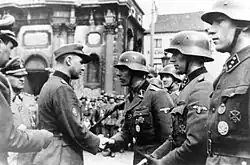 Photographie en noir et blanc de Léon Degrelle à Charleroi. Degrelle est à la gauche de l'image. En uniforme allemand et portant une casquette, il serre la main de l'un de ses hommes