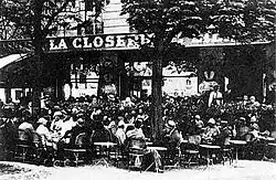 Vue en noir et blanc d'une terrasse de café bondée de consommateurs assis, au début du XXe siècle.