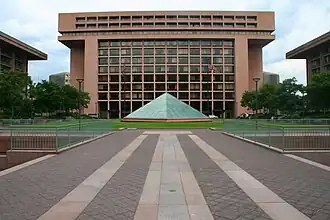 Vaste esplanade devant pyramide en verre et bâtiment aux formes géométriques et vitrages nombreux.