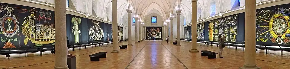 Photographie montrant des tapisseries à dominantes noires et or accrochées dans un salle voûtée d'ogives.