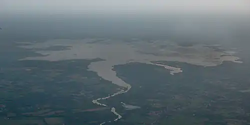 Plan large du lac pris d'avion ; le ciel est brumeux, on distingue les vastes zones de végétation flottante. Au premier plan, un paysage agricole et boisé, avec des zones d'habitation.