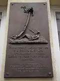 Photographie depuis le bas d'une plaque en métal sombre ; la partie supérieure étant une sculpture en relief, la partie inférieur un texte en hongrois sur plusieurs lignes