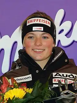 Photographie d’une jeune fille, blonde, avec un bandeau sur la tête, en buste, tenant un bouquet de fleurs et un trophée devant elle.