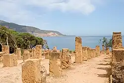 allée bordée de piliers rectangulaires en ruine, avec la mer en arrière-plan