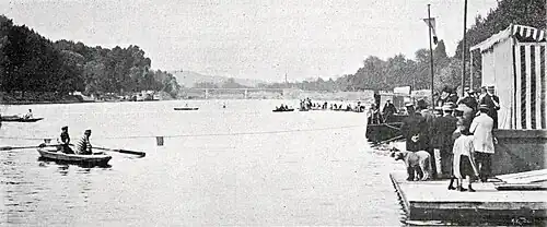 Photographie noir et blanc : des barques sur une large rivière ; une foule au bord de l'eau à droite.