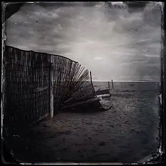 Photographie en noir et blanc représentant une plage déserte avec à gauche une clôture légère s'afaissant sur elle-même.
