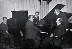 Un homme costaud et joufflu en costume, imposantes moustaches, qui joue du piano, entouré de quatre autres hommes moustachus en costume qui le regardent.