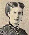 Half-length photographic portrait of a young lady with dark-colored hair swept back and wearing a high-necked, dark Victorian era satin dress with dark buttons