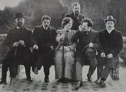 Image en noir et blanc montrant quatre hommes et une femme assis, et un autre homme debout derrière eux