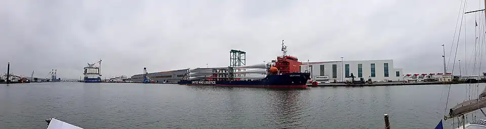 Livraison des pâles d'éoliennes en mer dans le port de Saint Nazaire. Au second plan, le navire Vestvind est à quai, chargé des pales d'éoliennes qui seront montées sur les turbines des éoliennes offshore. À l'arrière plan à gauche, le paquebot Celebrity Beyond en cours d'armement dans le bassin de Penhouët. À l'arrière plan à droite, l'usine de moteurs thermiques Man, et à l'extrême droite les turbines entreposées en attendant de repartir vers le champ en mer.
