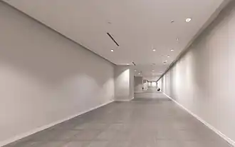 Long couloir et forte impression de perspective avec murs et plafond nus, avec quelqu'un assis par terre, seul au loin, à la station MRT Marina Bay, métro de Singapour. Juin 2018.