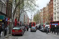 Une rue pavée bordée d'immeubles anciens rouges et blancs. Parmi la circulation routière se trouvent deux bus rouges à étage.