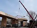 Photo couleur d'un pont en bois, circulaire et surélevé, en cours de construction. Au premier plan : deux voitures, un camion et une grue rouge. L'arrière-plan est constitué d'un ciel bleu nuageux.