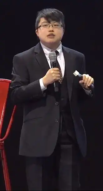 Personne aux cheveux courts portant des lunettes, portant un costume et s'adressant au microphone