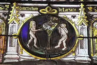 Médaillon de vitrail médiéval représentant la Tentation d'Adam et Eve, un homme et une femme sont nus autour d'un arbre portant des fruits et dans lequel il y a un serpent, la femme tient une pomme et l'homme a commencé à en manger une autre