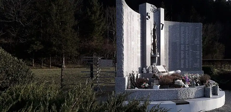 À droite, le monument aux morts en marbre ; derrière le monument, un grillage protège un tourniquet en métal.