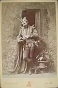photo recto : la basse Comte, en tenue de scène Méphistophélès, devant des décors, un coffret de bijoux à la main
