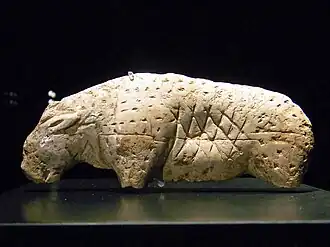 Photographie en couleurs sur fond noir d'une statuette en ivoire posée sur un socle noir et représentant un lion des cavernes aux pattes amputées.